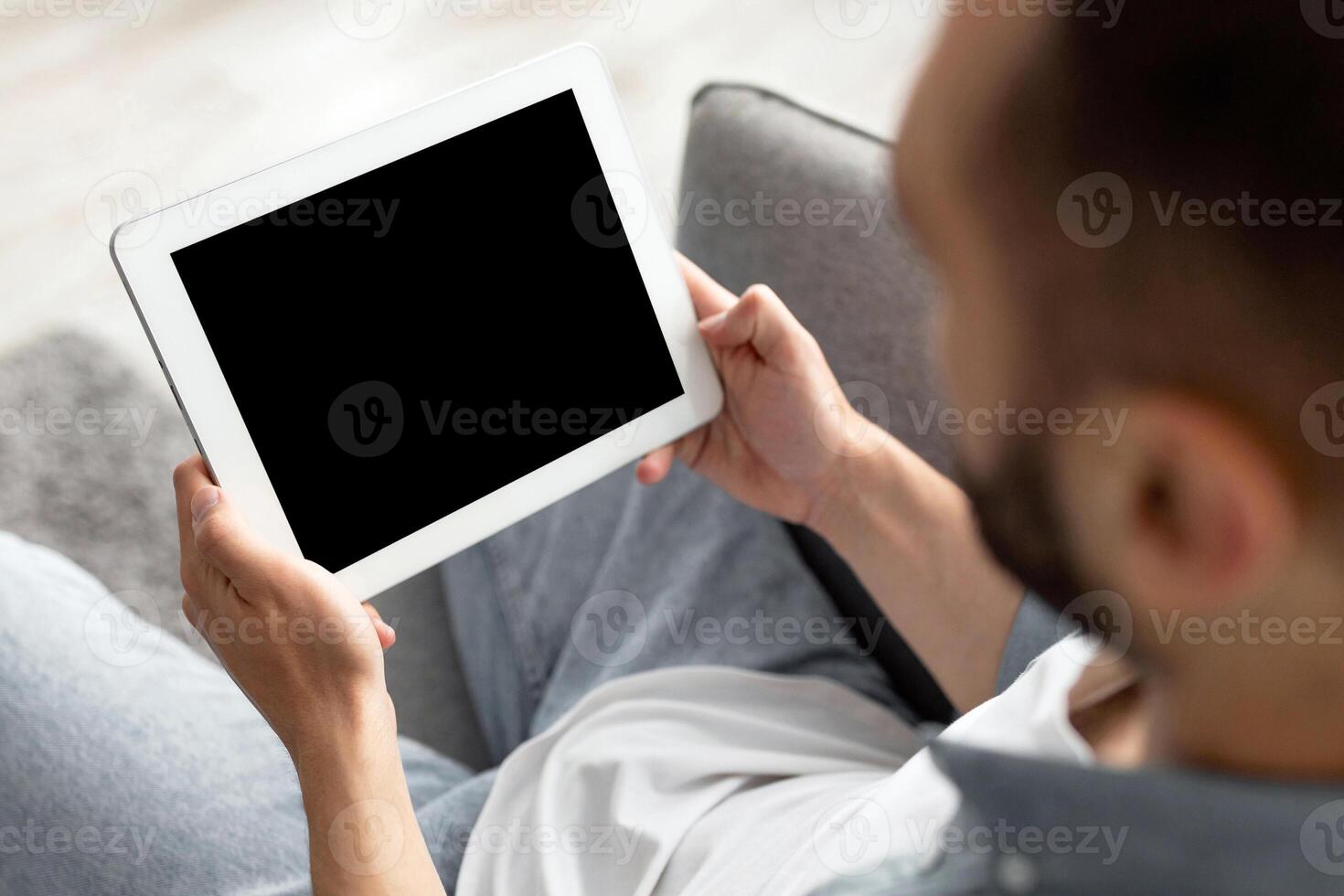 Unrecognizable young guy working on tablet computer with empty screen at home office, mockup for website or app design. Closeup of millennial man holding touch pad, space for advertisement photo