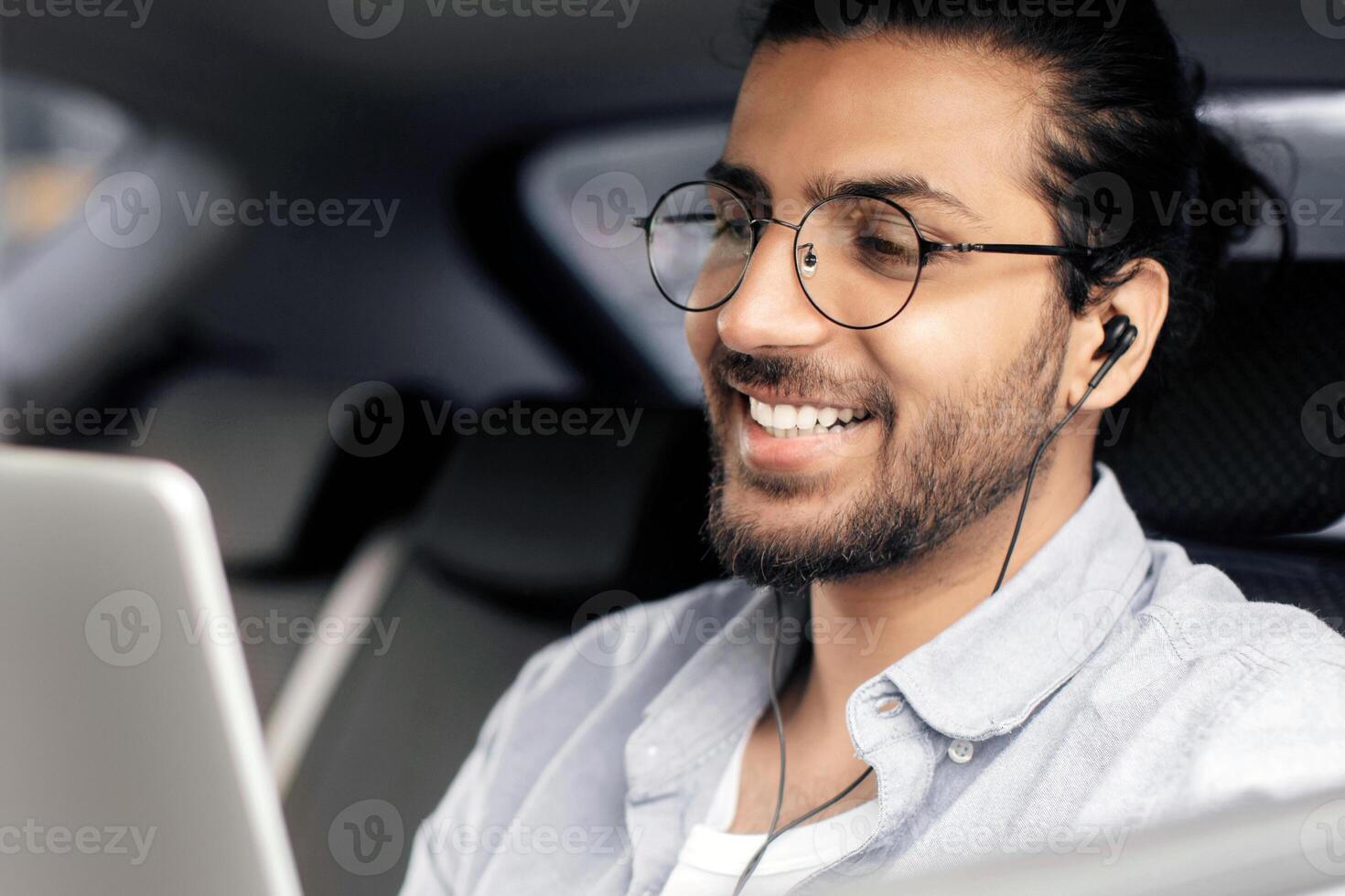 Smiling arab guy sitting at car back seat, listening to music while going somewhere by transportation service, using earphones and mobile application on digital tablet, copy space, closeup photo
