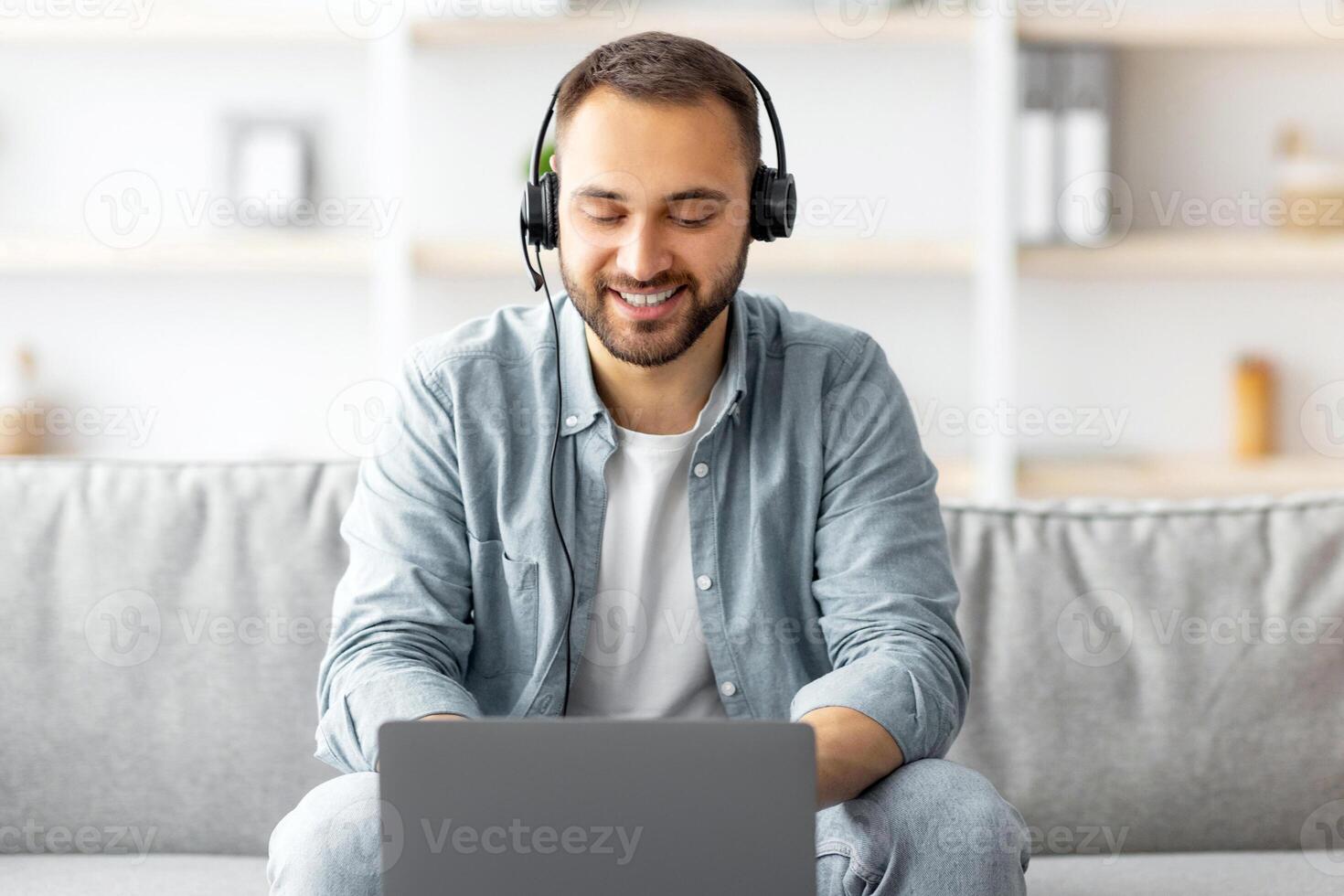Smiling young Caucasian guy in headset making call on laptop at home, talking to webcam. Millennial man communicating online , participating in web conference or remote meeting photo