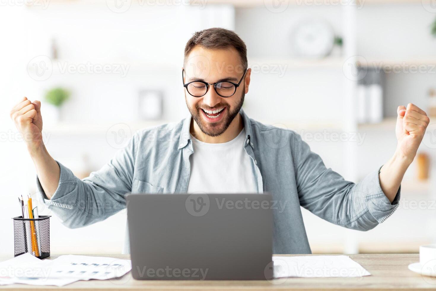Young Caucasian guy gesturing YES in front of laptop, celebrating success at home office. Millennial man working with portable pc, making great deal, signing online contract, excited over achievement photo