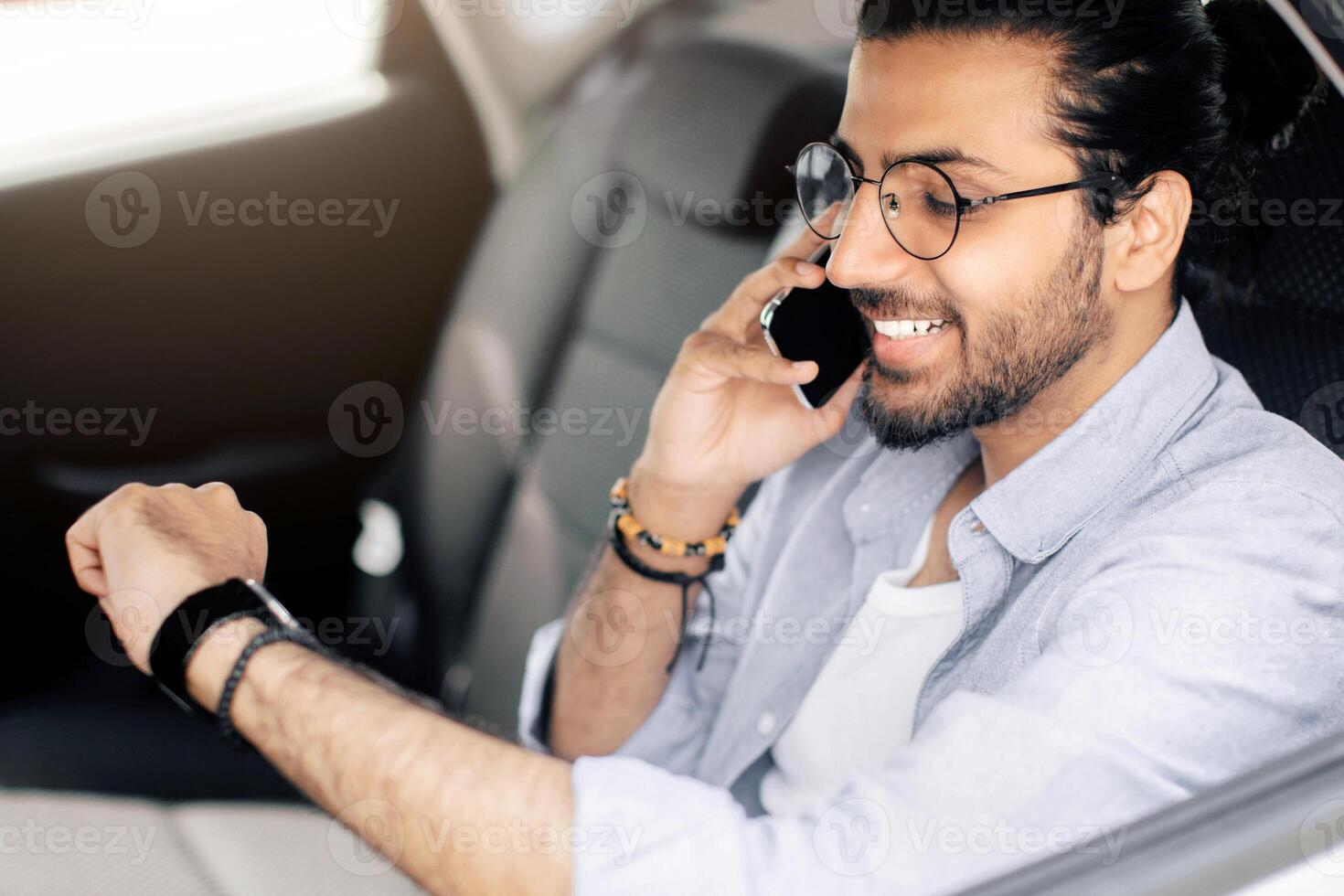Happy young indian man in casual outfit sitting at car back seat, having phone conversation, checking time, going to airport by taxi, middle-eastern guy taking cab, copy space photo