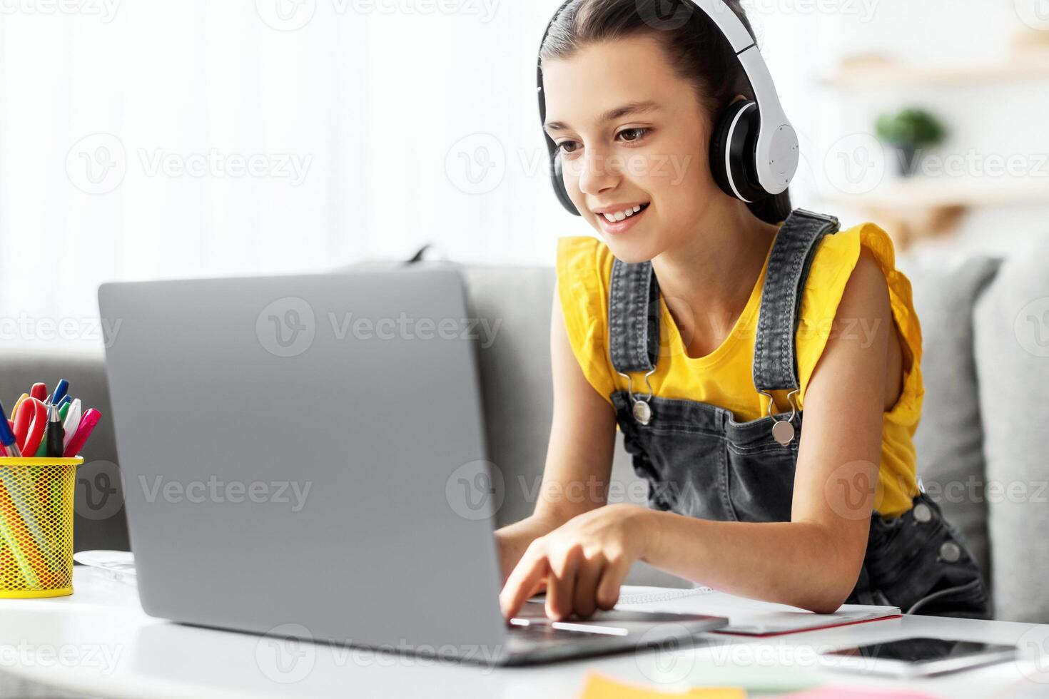 Kids And Technology Concept. Portrait of smiling girl sitting at table and using laptop computer, wearing wireless headphones, watching online course or movie, playing online game, free copy space photo