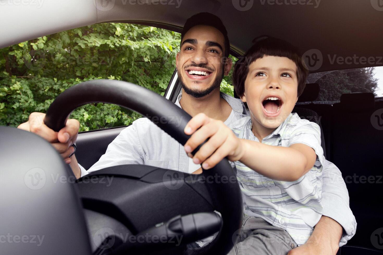 Portrait Of Excited Little Boy Driving Car With His Arab Dad, Holding Steering Wheel And Laughing, Caring Middle Eastern Father Teaching Son How To Drive, Enjoying Road Trip In Modern Vehicle photo