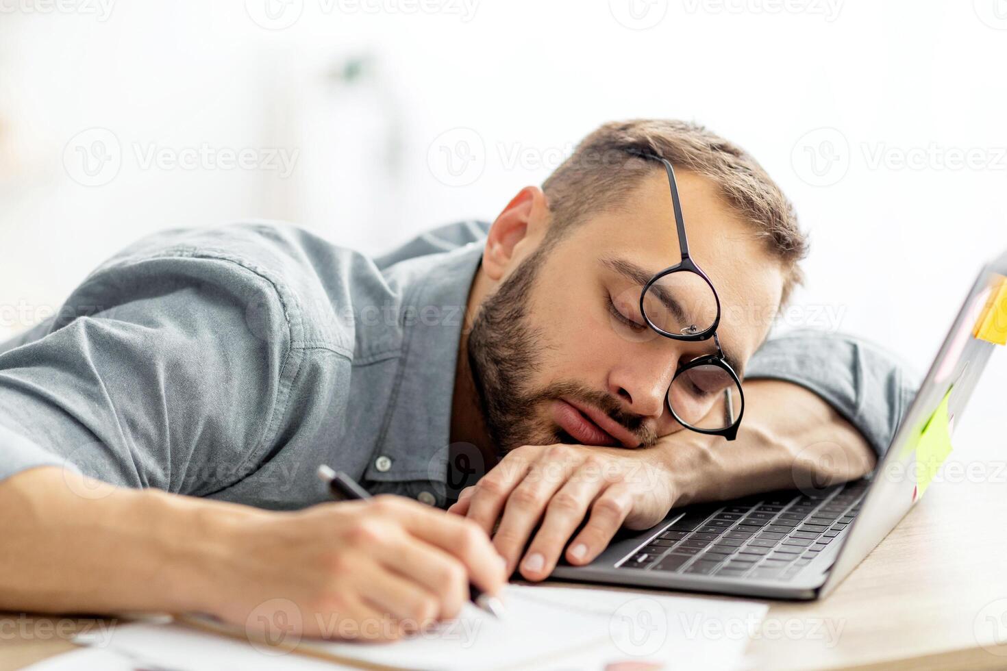Exhausted millennial man sleeping on his office desk, next to laptop and documents, tired of overworking. Young male workaholic suffering from chronic fatigue at workplace photo
