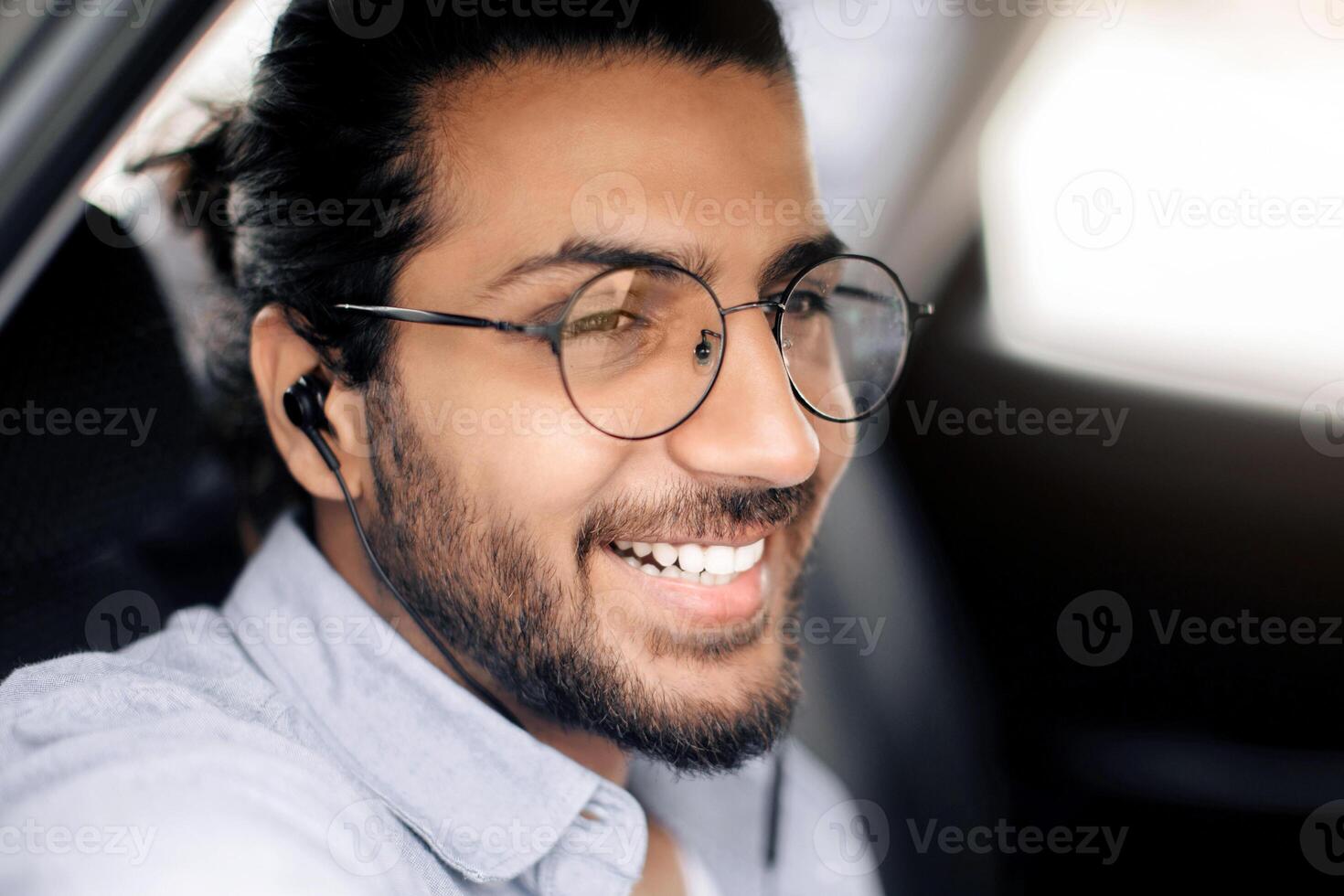 Closeup of middle-eastern long-haired guy with glasses listening to music in car, positive arab man with earphones looking through window and smiling, travelling by car, using transportation service photo