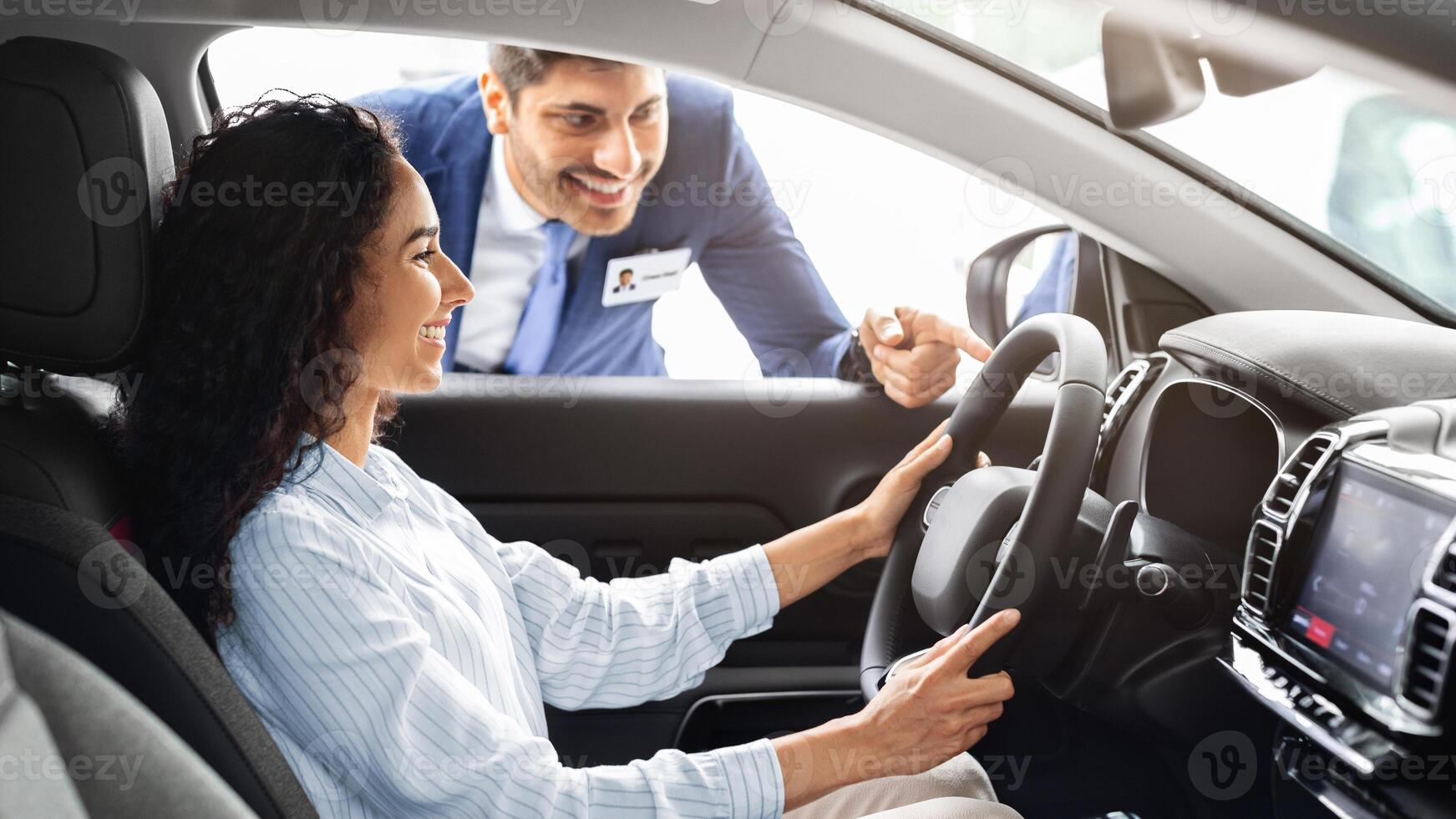 Young business lady buying new luxury car in new showroom, happy brunette woman sitting in nice automobile, holding hands in steering wheel, having conversation with sales assistant, copy space photo