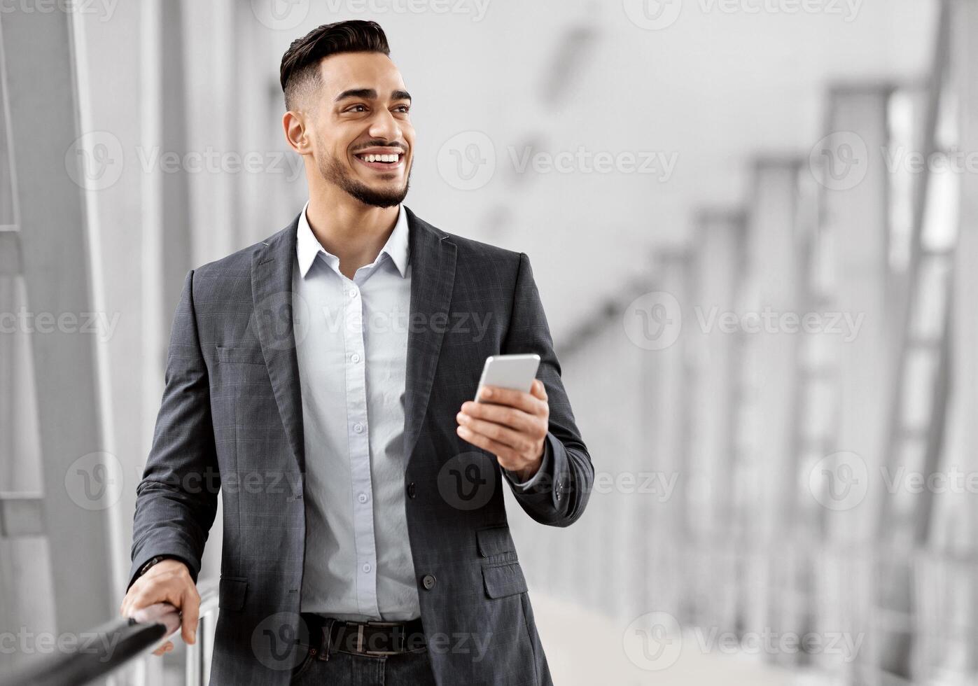 Handsome Middle Eastern Man With Smartphone In Hands Waiting In Airport Terminal, Smiling Young Arab Businessman Holding Mobile Phone And Looking Away, Enjoying Online Communication, Copy Space photo