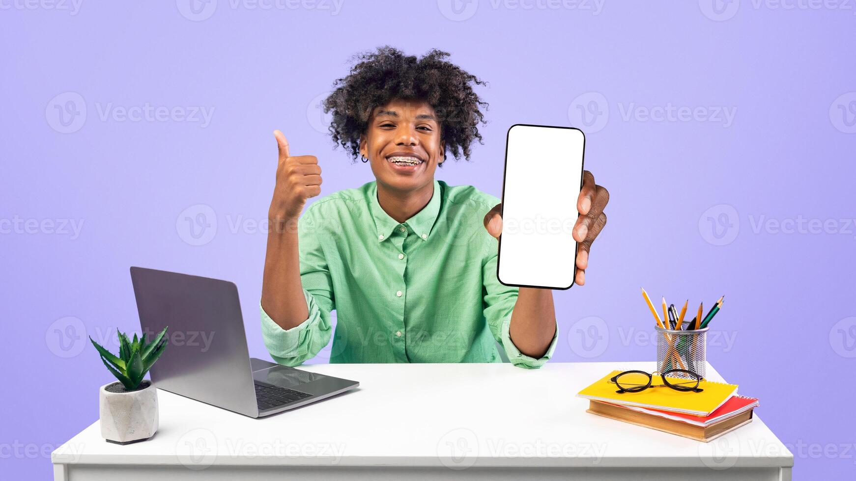 Cheerful black student guy holding smartphone with blank screen and showing thumb up, sitting at table over blue background. Teen recommending website for learning online photo