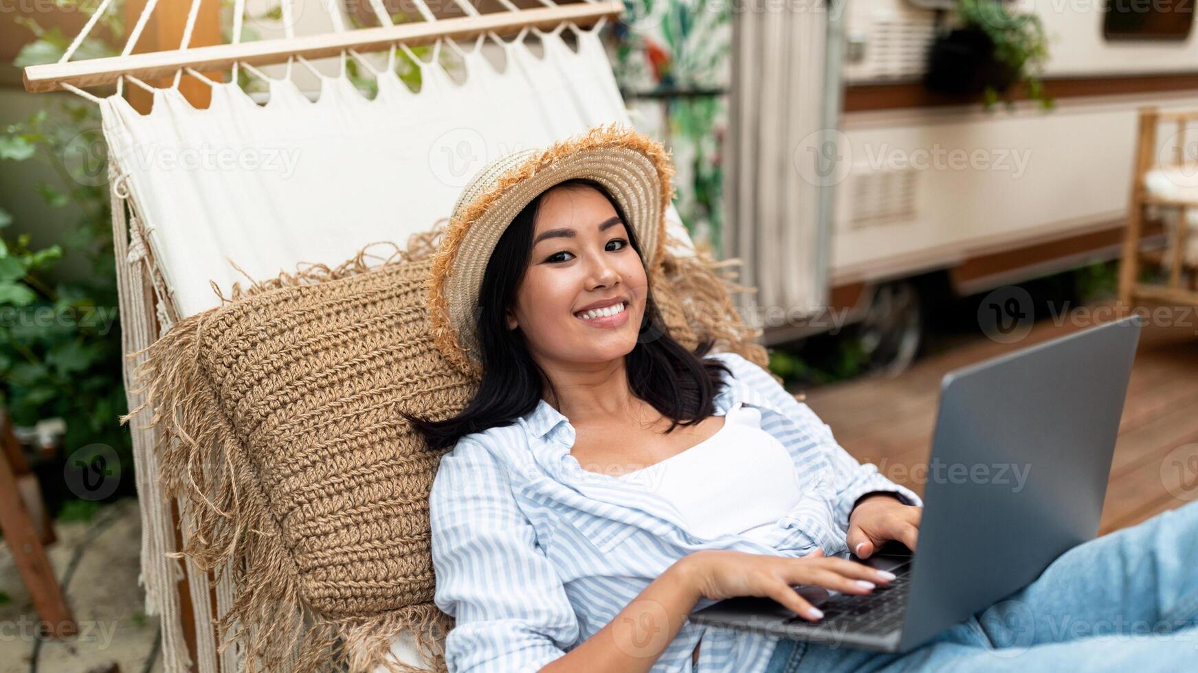 Joyful Asian lady using laptop computer while lying in hammock near RV on autumn camping trip. Cheery young woman working or studying online, browsing web outdoors on weekend photo