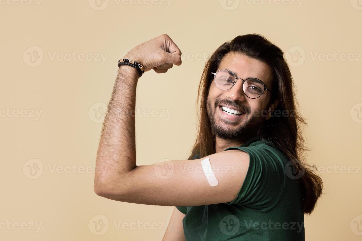 Handsome long-haired middle-eastern guy showing biceps with medical plaster on, got vaccinated against COVID-19 while pandemic, feeling strong and positive, beige studio background photo