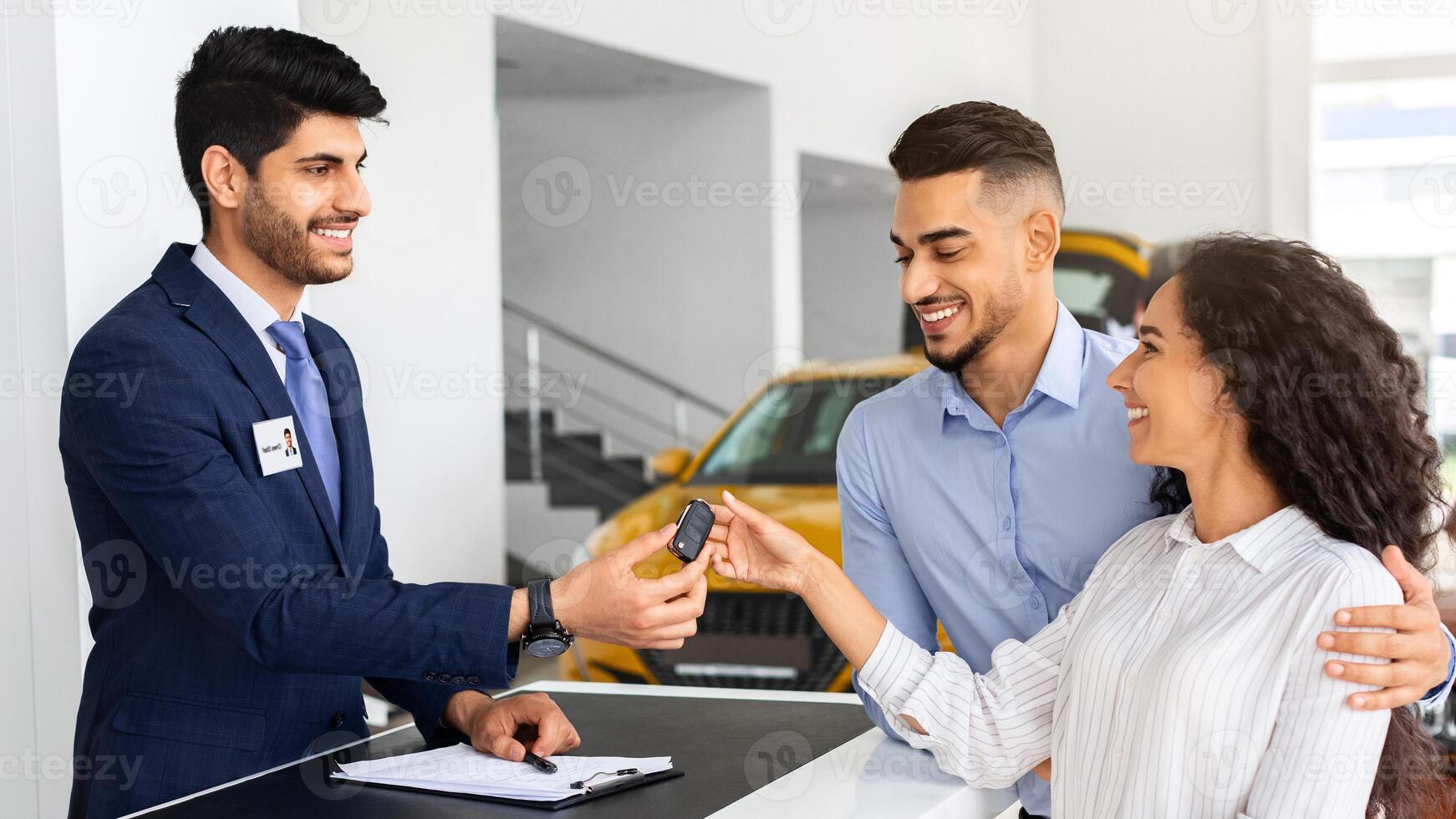Happy middle-eastern couple buying car in huge showroom, taking automobile key from young handsome salesman, side view. cheerful arab man and woman purchasing beautiful auto, hugging and smiling photo