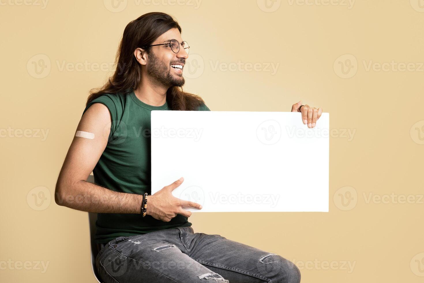 Cheerful middle-eastern vaccinated man with empty board posing on beige studio background, looking at copy space and smiling. Happy young arab guy recommending vaccination while COVID-19 pandemic photo