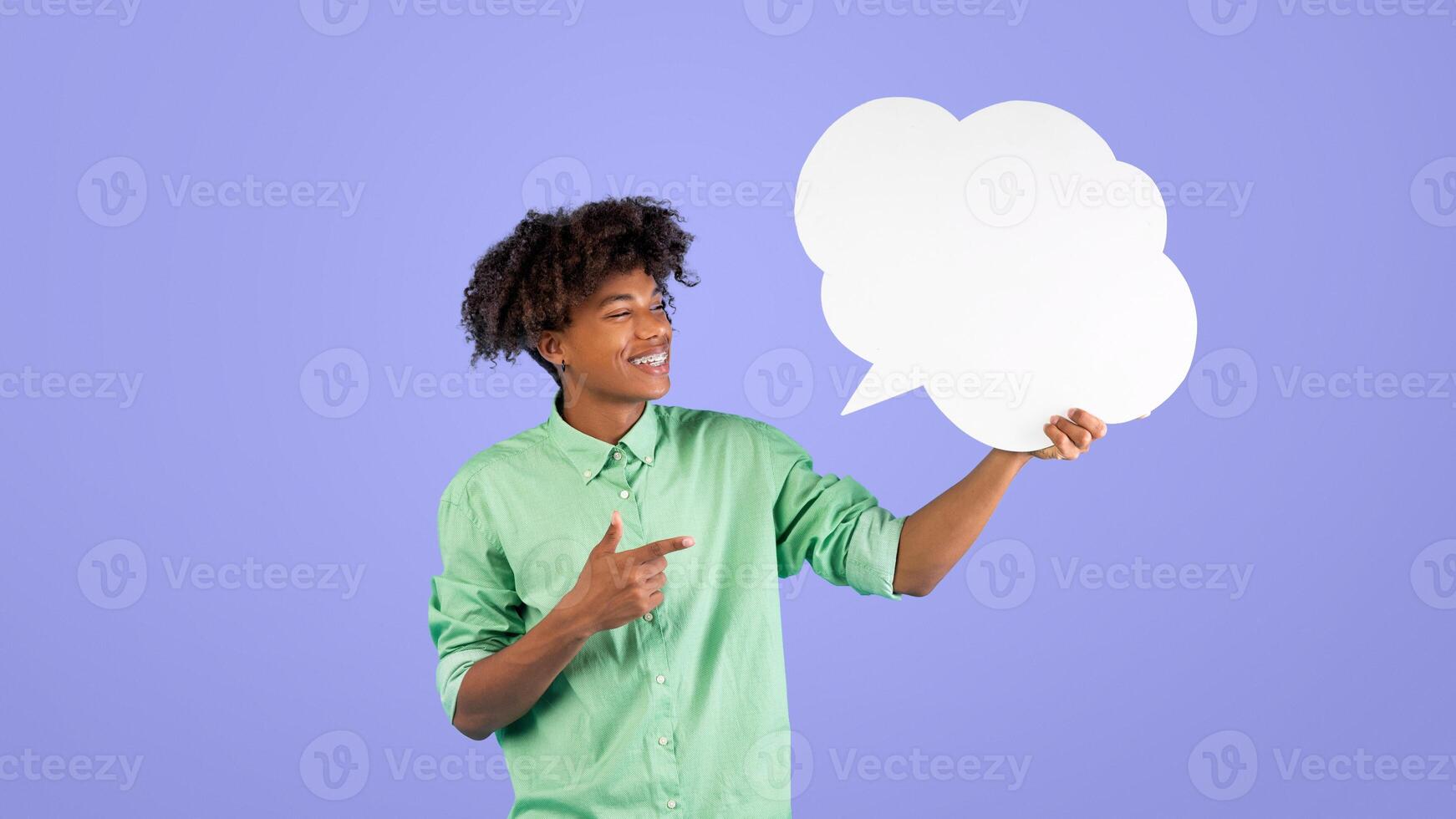 Cool african american guy pointing at empty speech bubble and smiling, standing over blue studio background, mockup for design. Happy teenager with blank word cloud, copy space photo