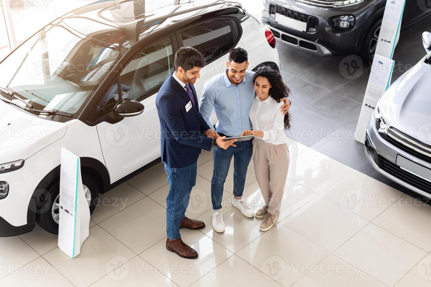 Top view of cheerful middle-eastern young couple having conversation with good-looking sales assistant at auto showroom, checking papers or car specification, full length photo