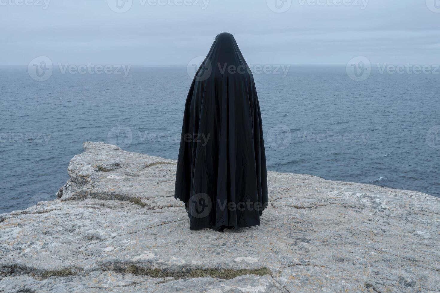 Mysterious figure in black robe overlooking the ocean from a cliff photo