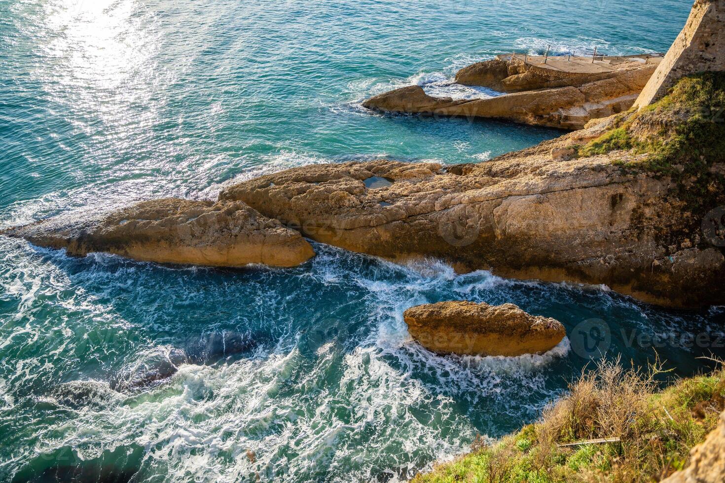 View of a sunlit rocky coastline with the sea shimmering under a clear blue sky in Ulcinj, Montenegro. High quality photo
