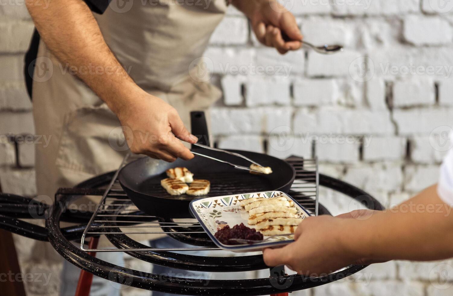Cooking grilled cheese. Chef hands holding tongs and putting pieces of cheese on plate. Waiter holds tray with soft traditional Cypriot pickled haloumi and jam. photo