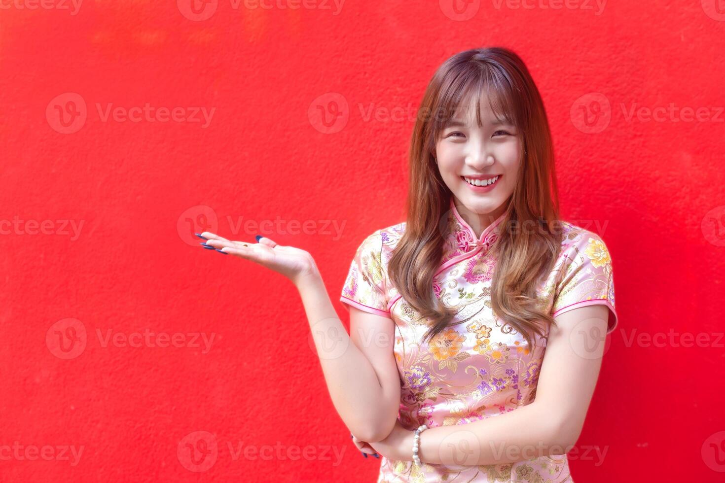 Young asian beautiful confident woman in pink dress stands is pointing hand to present something while isolated red background in Chinese new year theme. photo