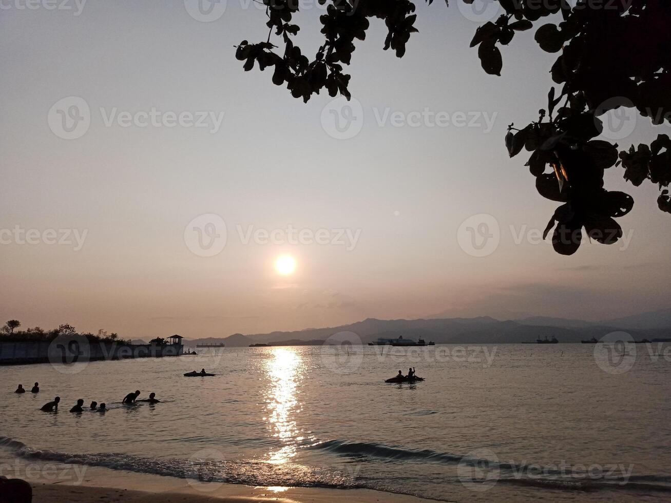 A serene sunset over a calm ocean with silhouettes of people swimming and boats sailing photo