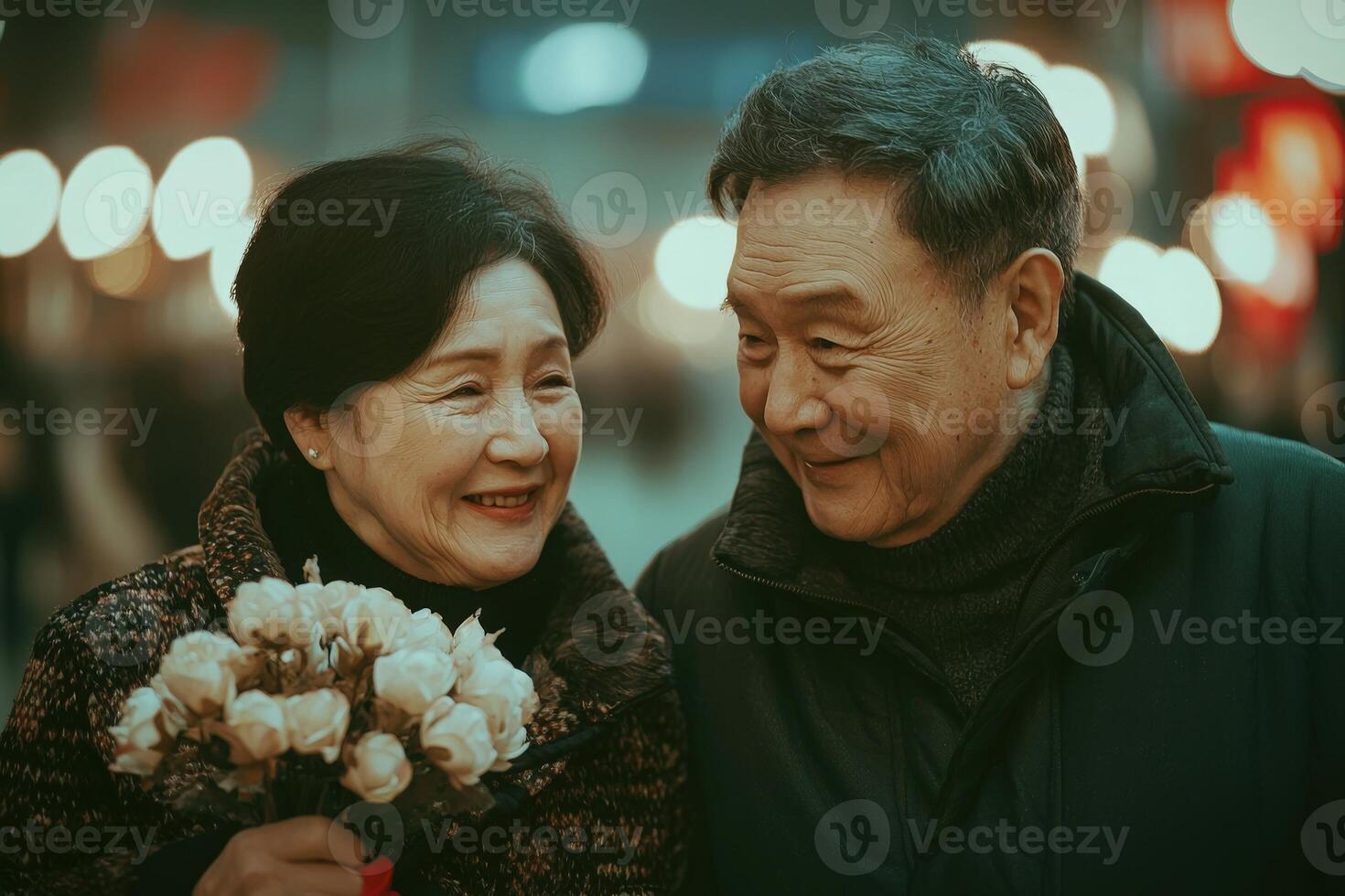 An elderly couple smiles at each other while holding a bouquet of flowers during a lively evening. Bright lights and a warm atmosphere enhance their happy moment together. photo