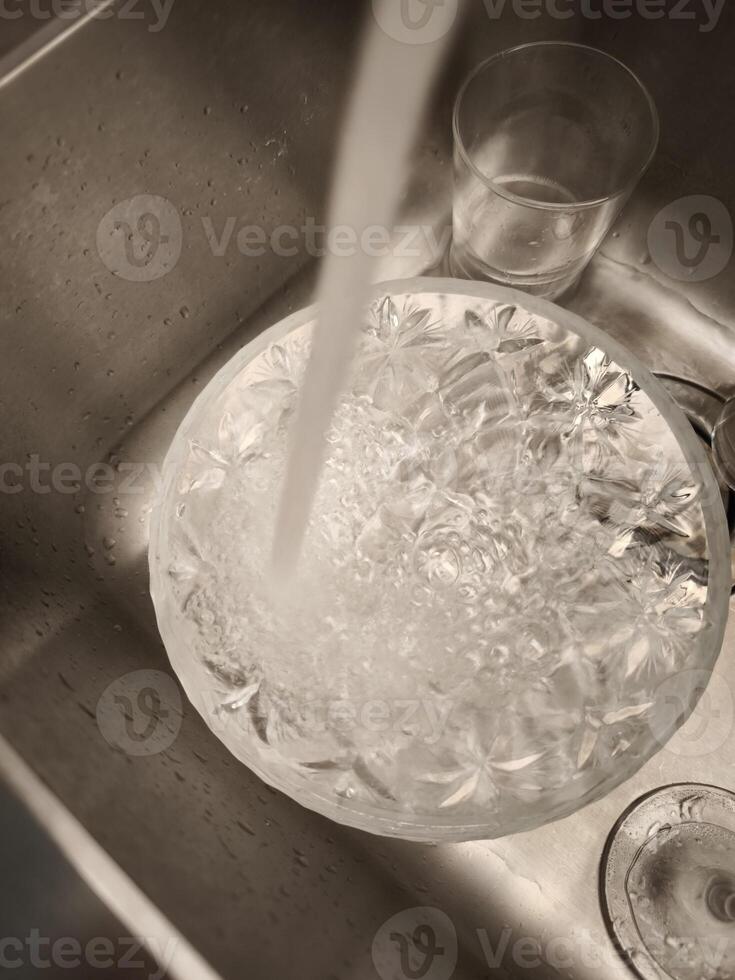 Crystal bowl filling with tap water in a stainless steel kitchen sink, with a drinking glass nearby, in prague, precious resource, take care of it World Water Day photo