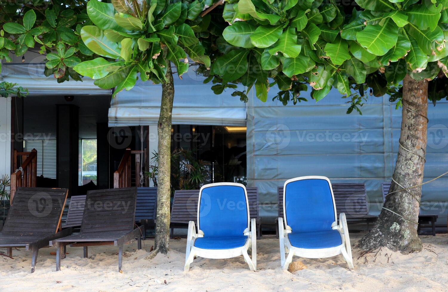 Two blue sun loungers and wooden deck chairs sit under lush green trees on a sandy beach near a beach hotel, evoking a sense of relaxation, vacation and tropical tranquility. photo