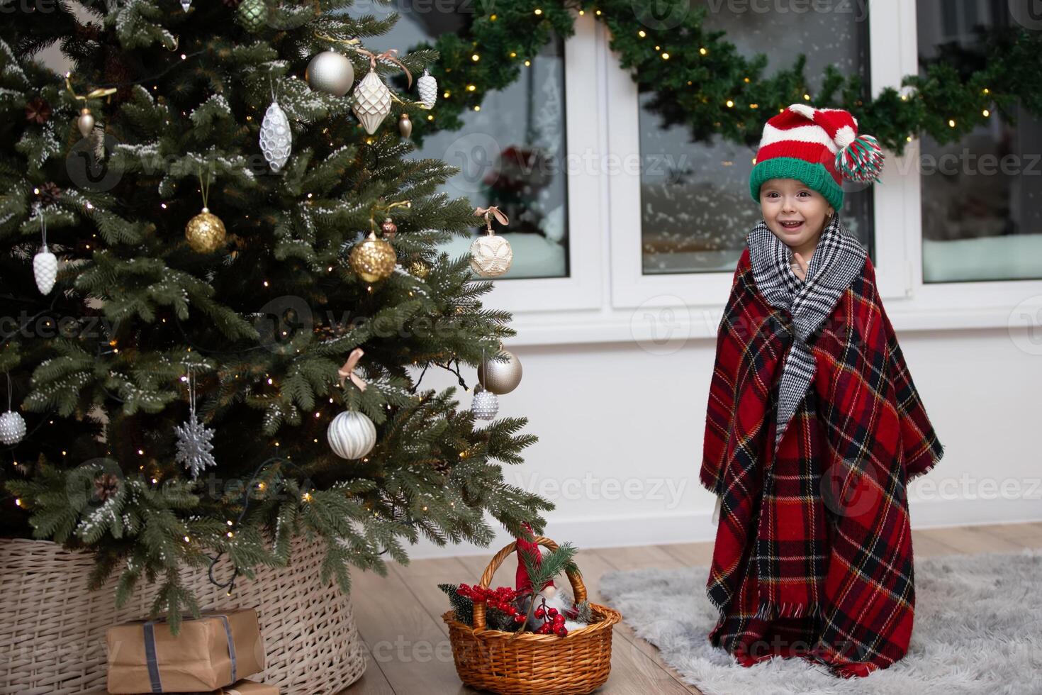 A happy little girl in a Santa Claus hat is wrapped in a warm plaid blanket by the Christmas tree. photo