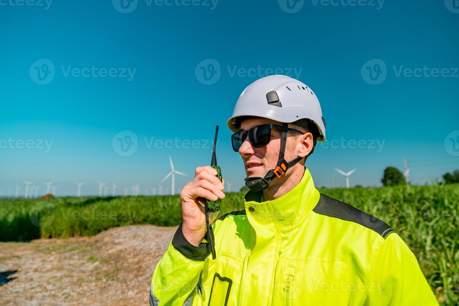 Worker in bright gear communicates with walkie-talkie in a field with wind turbines under clear skies photo