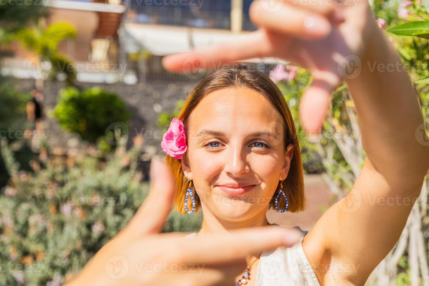 A woman with a pink flower in her hair is smiling and making a square with her fingers, summer vibes photo