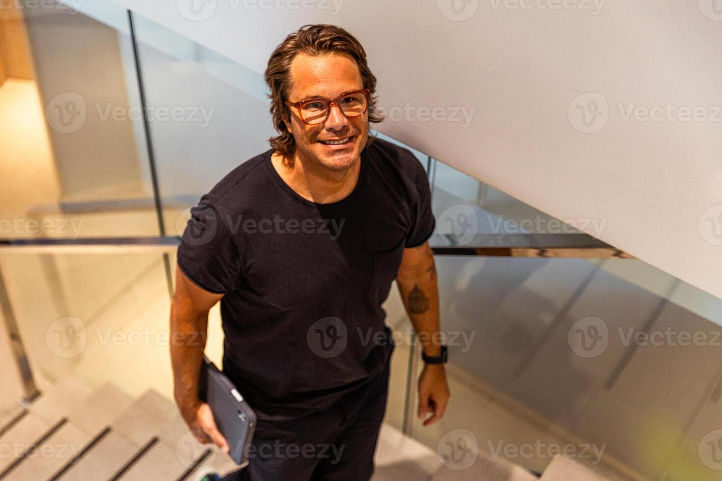 A man wearing glasses and a black shirt is smiling and holding a tablet, photo from above