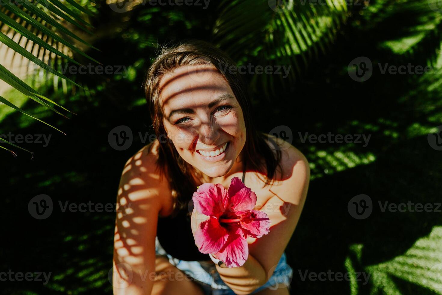 Smiling woman holding a pink flower while sitting on a grass field. Shadow of leafs on her face photo