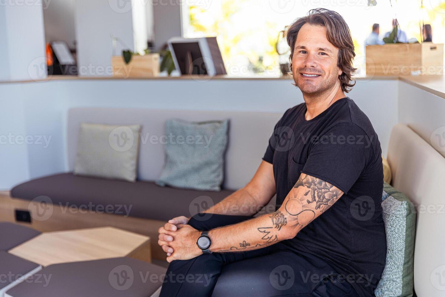 A man with tattoos on his arm sits on a couch smiling in a room photo