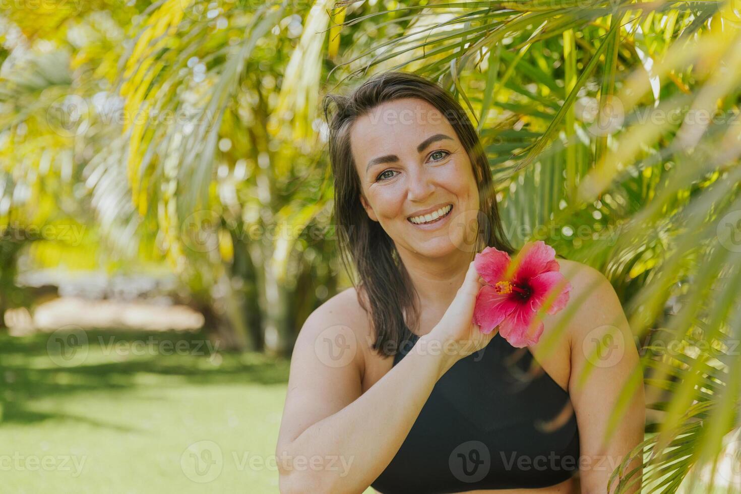 A woman is posing with a pink flower in her hand smiling and enjoying nature photo