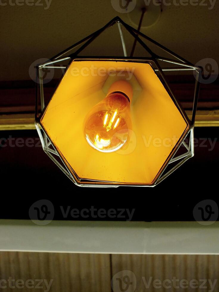 Light bulb is lit up in a hexagonal shaped lamp. The light is yellow and the lamp is hanging from the ceiling photo