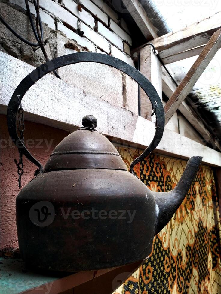 Black tea kettle sits on a wooden shelf. The kettle is old and rusted, and it has a chain hanging from it. The scene is somewhat eerie and gives off a sense of nostalgia photo