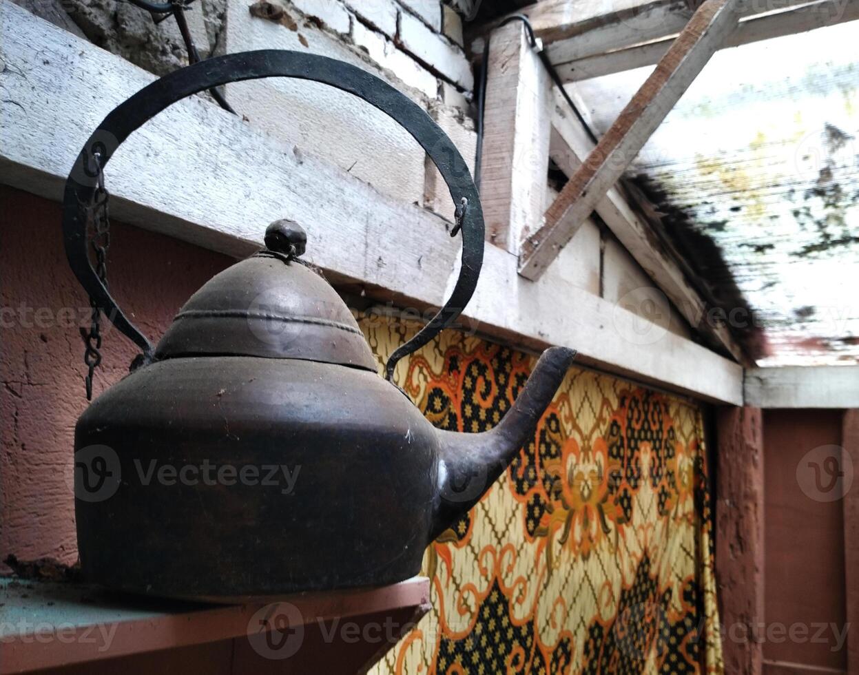 Black tea kettle sits on a wooden shelf. The kettle is old and rusted, and it has a chain hanging from it. photo