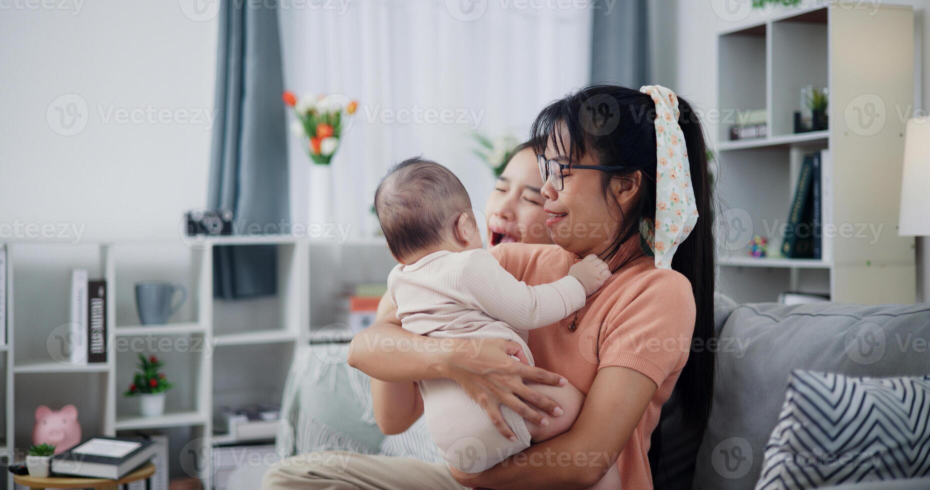 Asian mother sit on the sofa holding her little son in arms. The older sister leaned in to tease younger brother and made funny faces. photo
