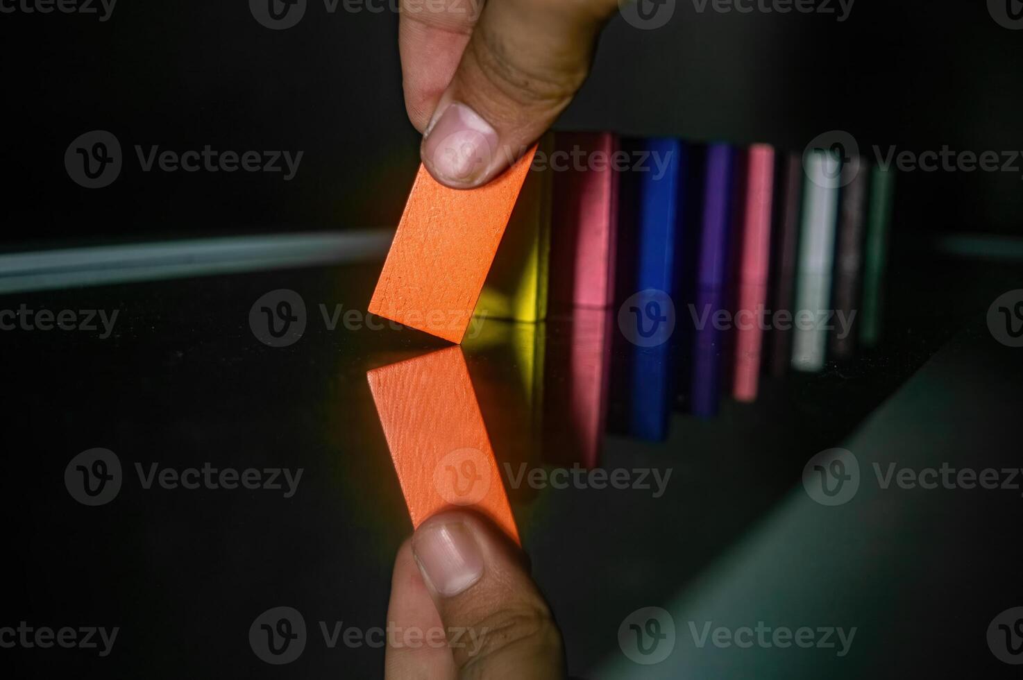 A hand is seen tipping an orange domino, initiating a chain reaction with other colorful dominoes lined up on a reflective surface. The image captures the moment of action and reflection photo