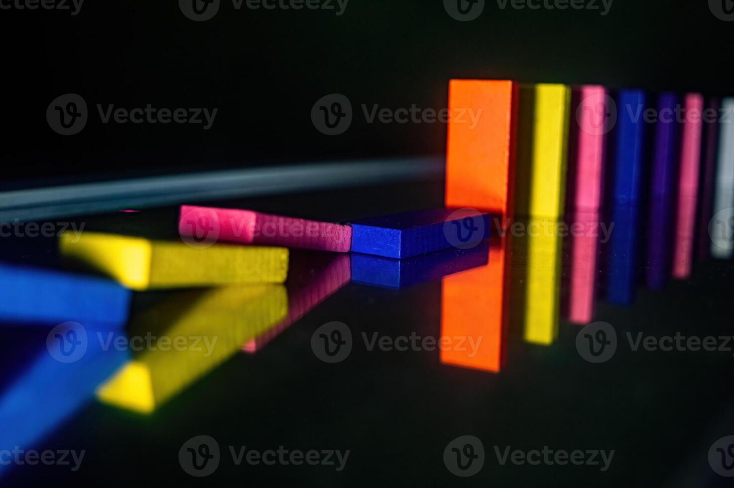Colorful dominoes in a line, with some fallen over, reflecting on a shiny surface. The vibrant colors and reflections create a visually striking and dynamic composition photo