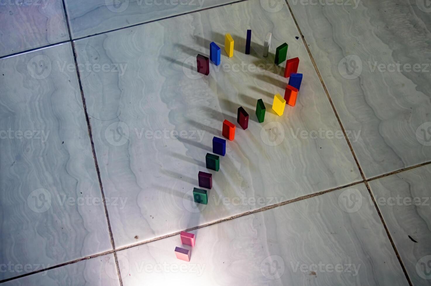 Colorful dominoes arranged in a question mark shape on a tiled floor, casting shadows. The arrangement and lighting create an intriguing visual effect photo