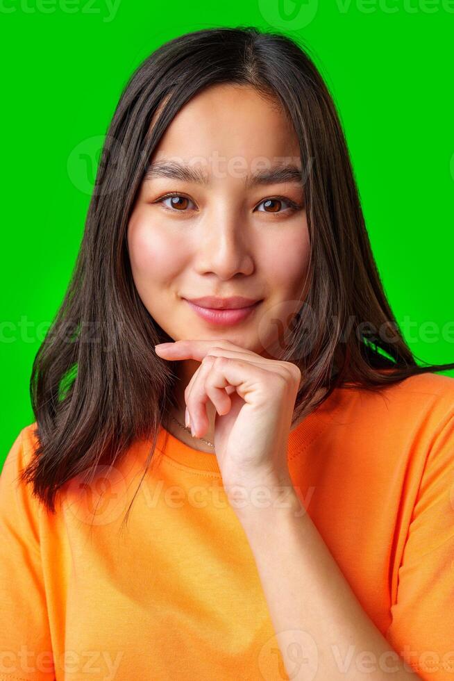 Young woman with long dark hair smiles while thoughtfully resting her chin on her hand in front of a vibrant green background photo