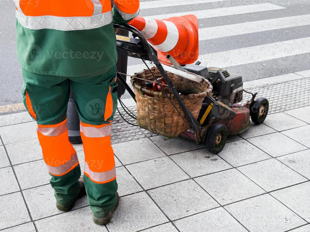 Worker in hi vis gear with lawn mower and traffic cone on urban sidewalk photo