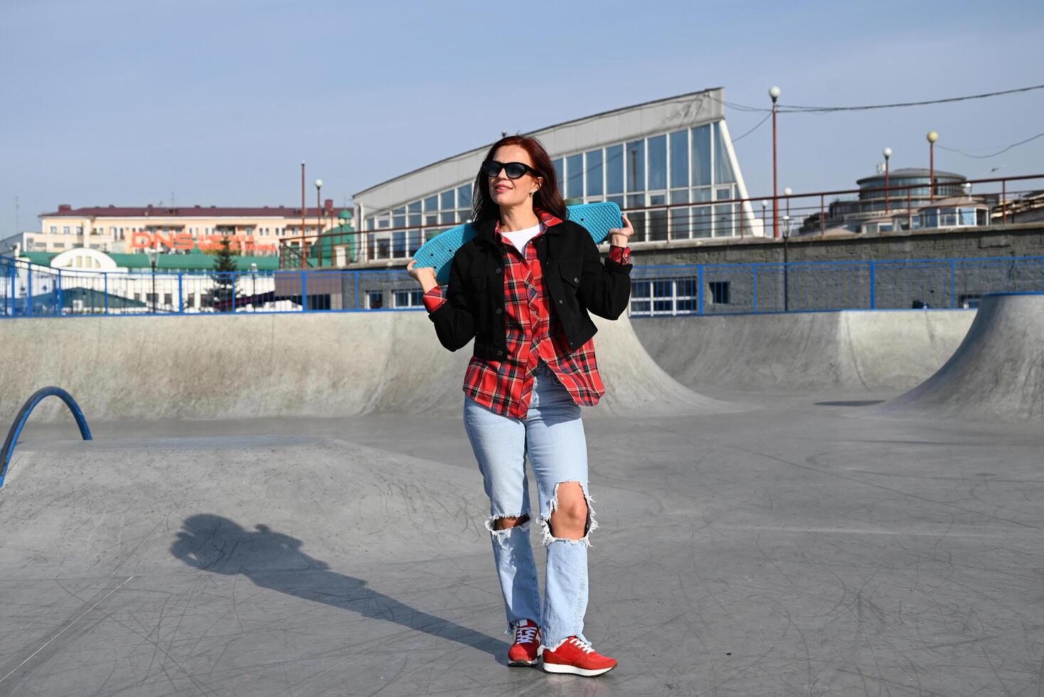 The photograph captures a casual yet stylish look of a person with a skateboard, set against a plain gray wall, highlighting modern, youth-oriented style photo
