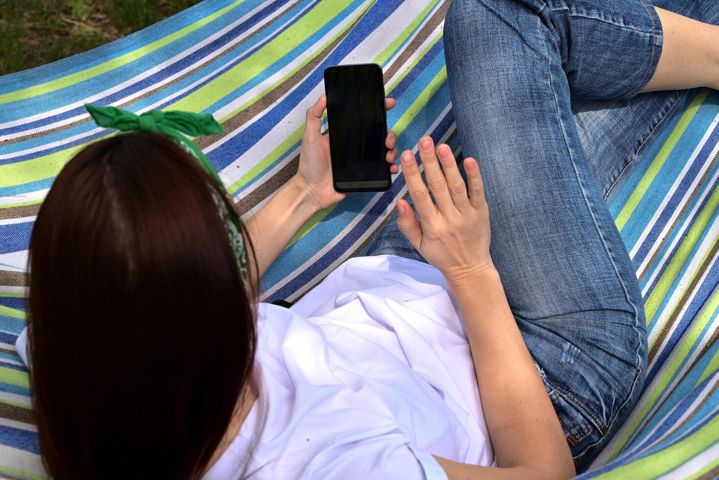 On a wonderful day with a positive attitude A young happy woman sitting in a park on the green grass in nature with a phone in her hands in casual clothes and chatting by link, the concept photo