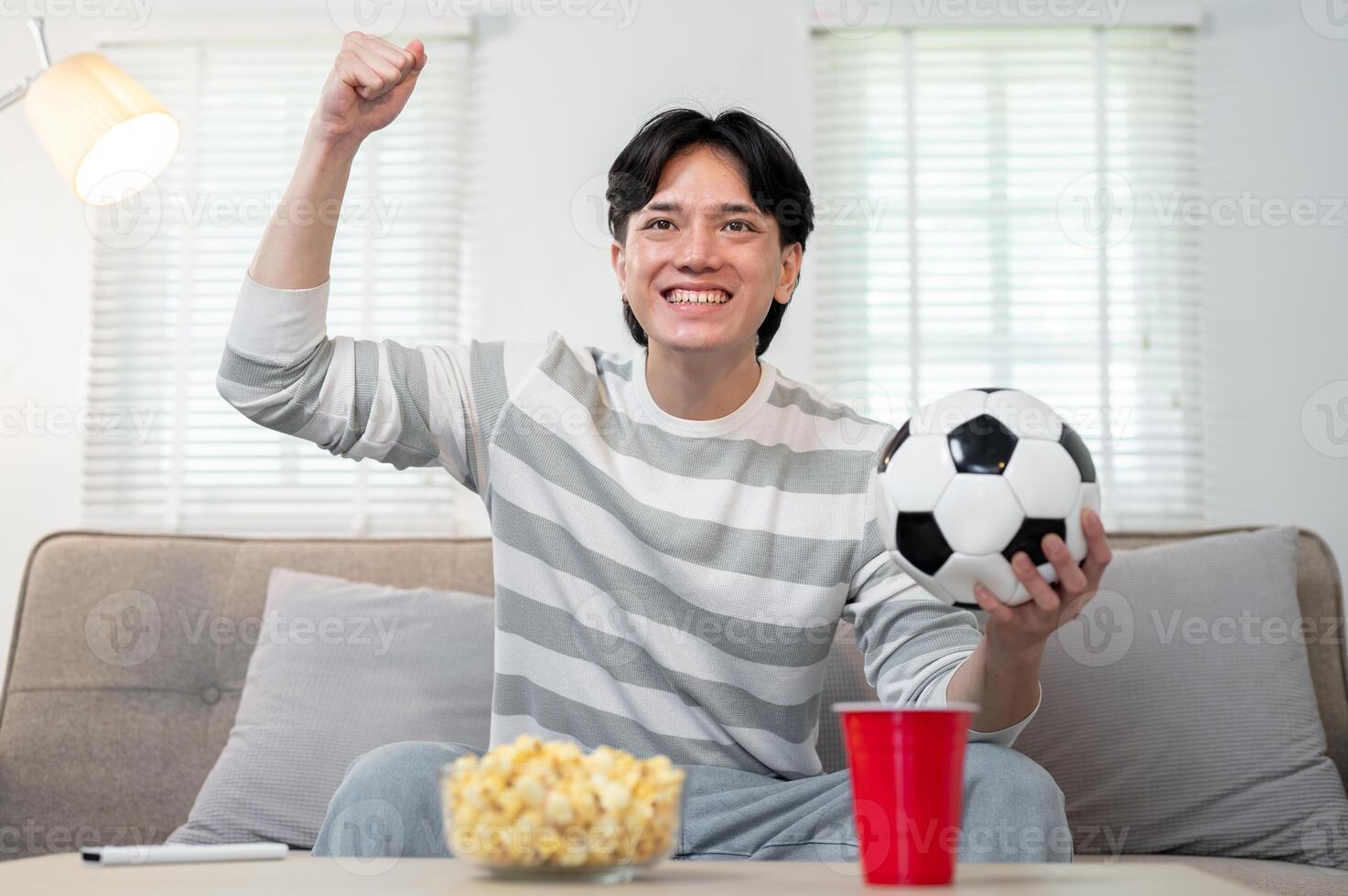An excited young Asian man is watching a football match on TV at home, cheering for his team. photo
