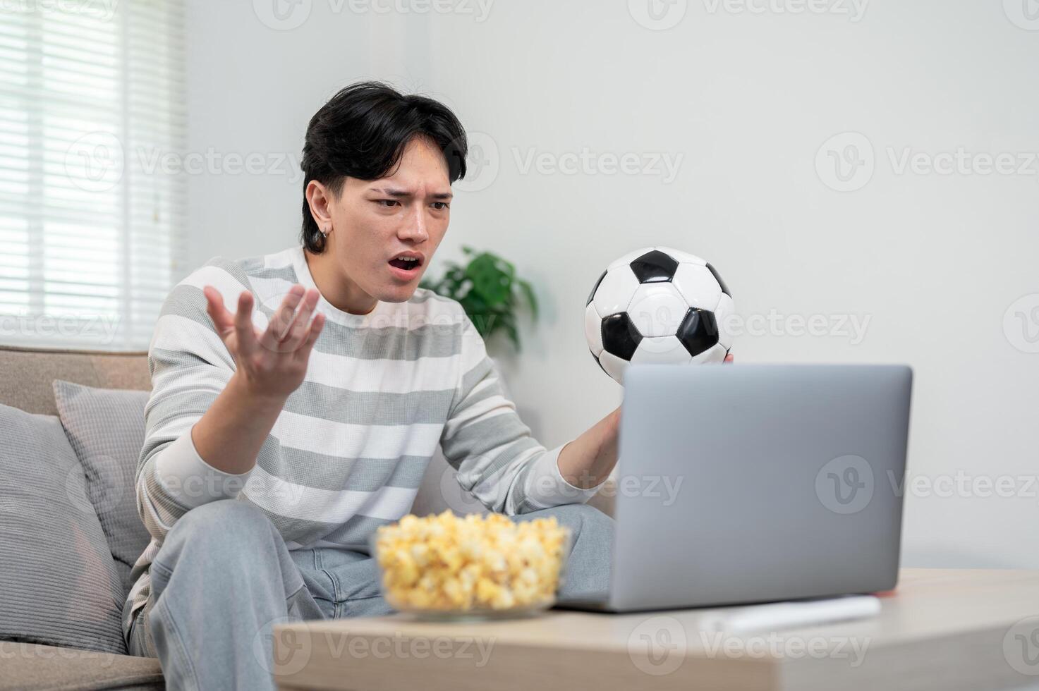 A shocked Asian man is watching his favorite football team lose to another team via his laptop. photo