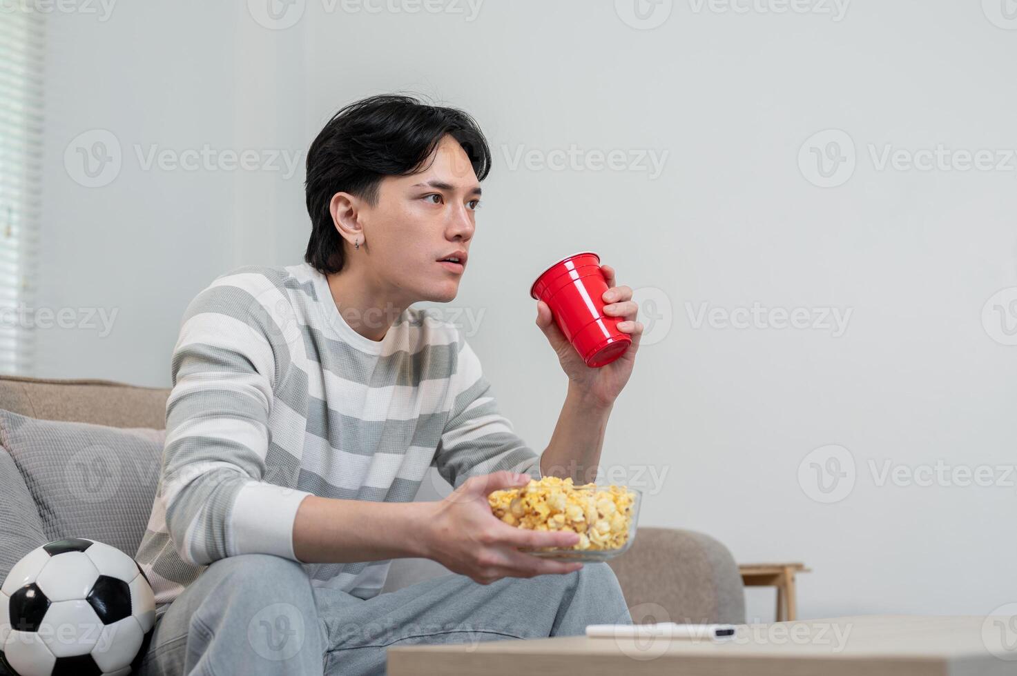 An excited Asian man, focused on his favorite football team's match on TV, sitting on the sofa. photo