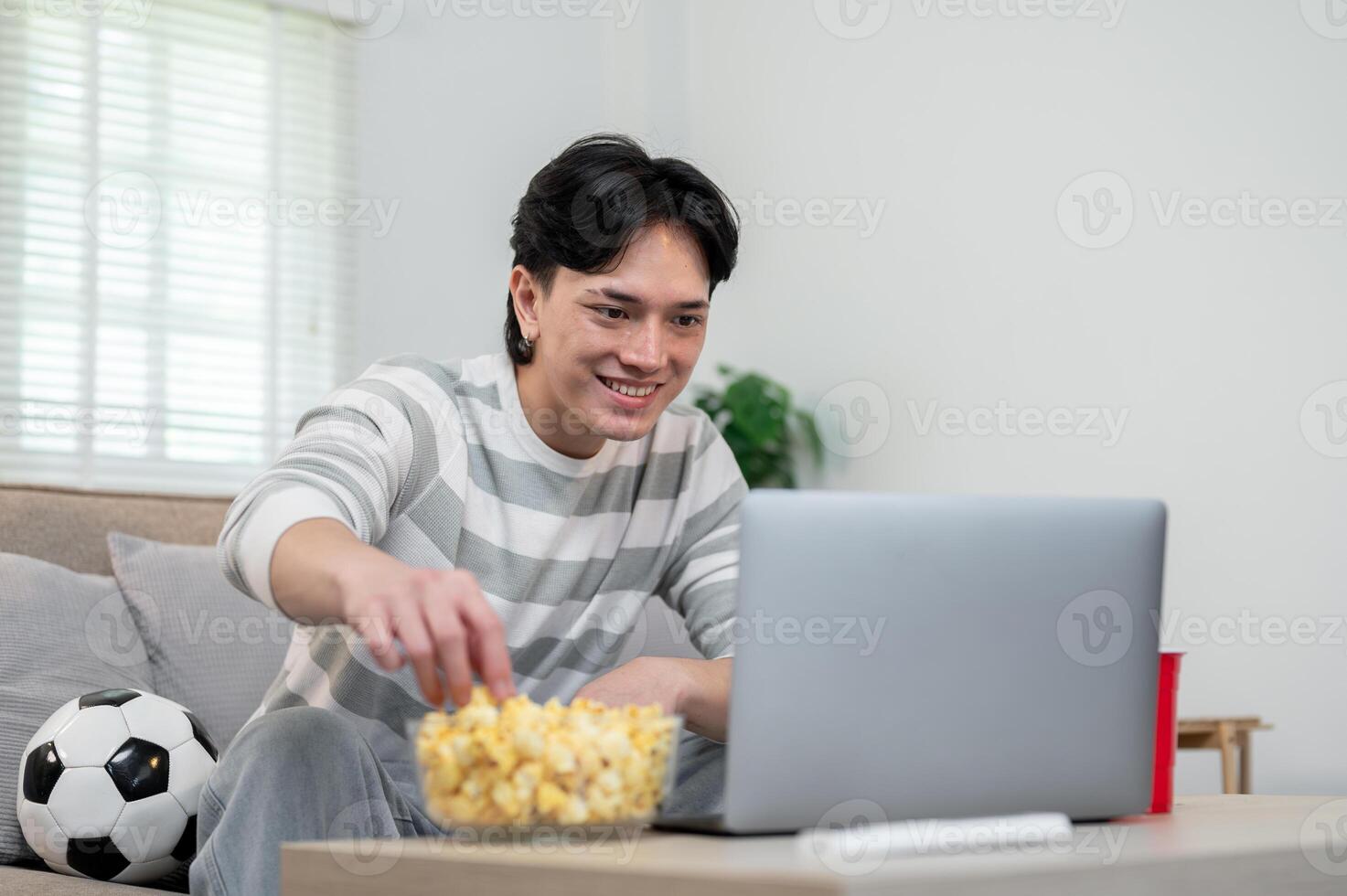A relaxed Asian man enjoys eating popcorn while watching a football match on his laptop at home. photo