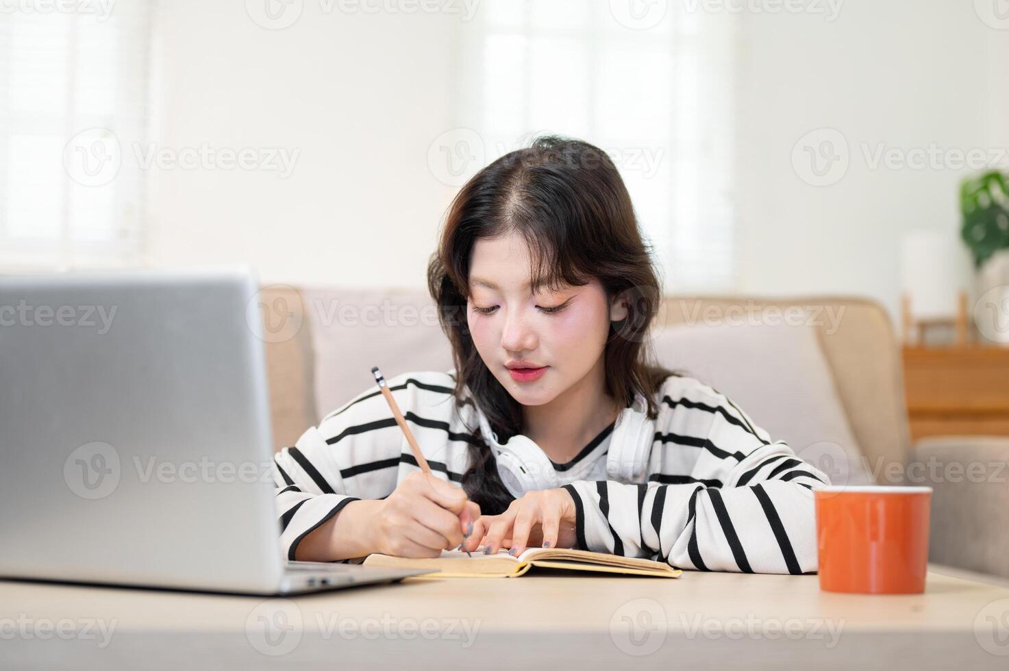 A beautiful Asian female student is studying online at home, taking notes from a lecture in her book photo
