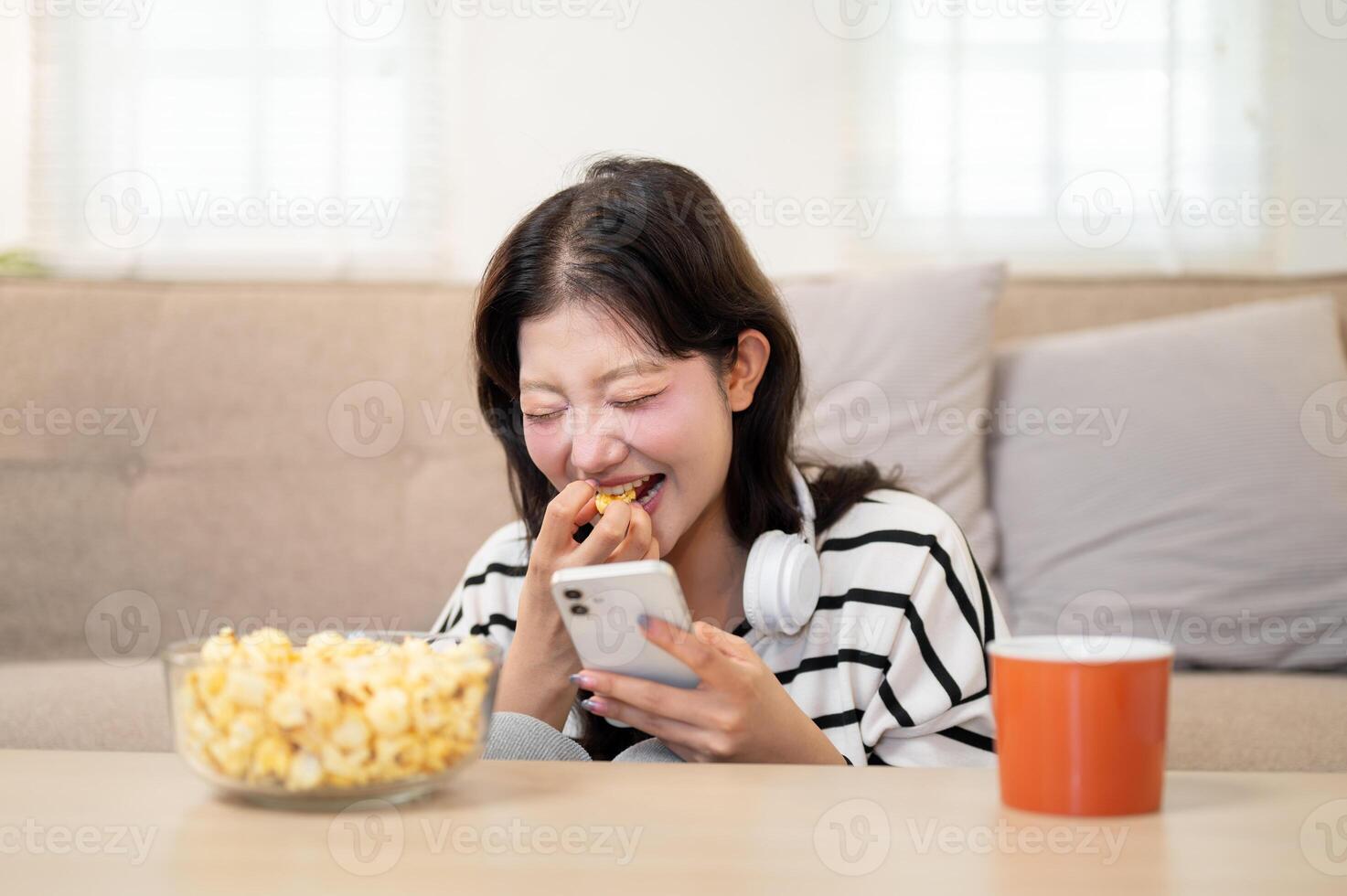 Cheerful Asian woman is laughing at the chat she's reading on her smartphone while enjoying popcorn photo